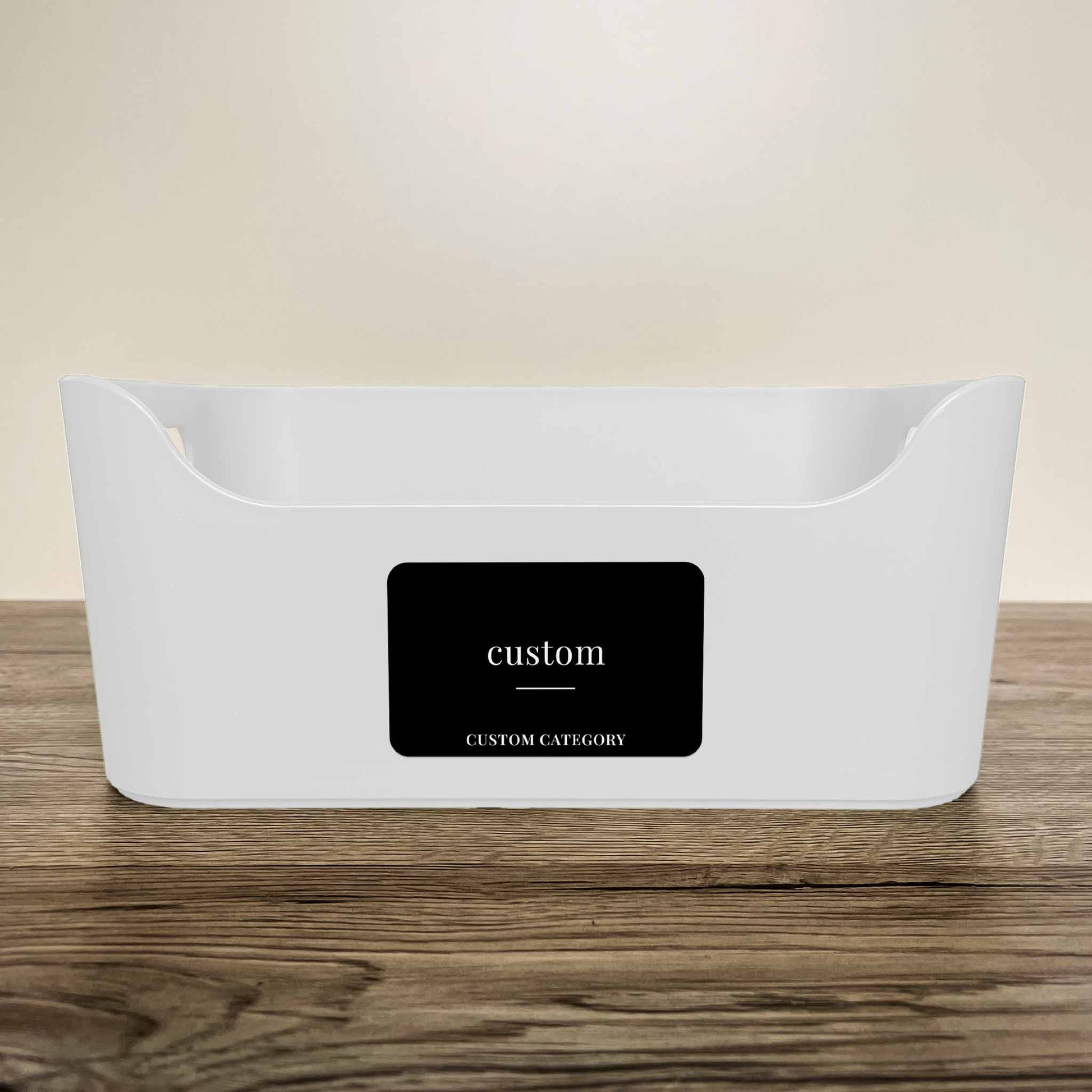 A white plastic storage basket with a customisable minimalist label in black colour on the front, displayed on a wooden surface against a neutral beige background.