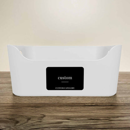 A white plastic storage basket with a customisable minimalist label in black colour on the front, displayed on a wooden surface against a neutral beige background.
