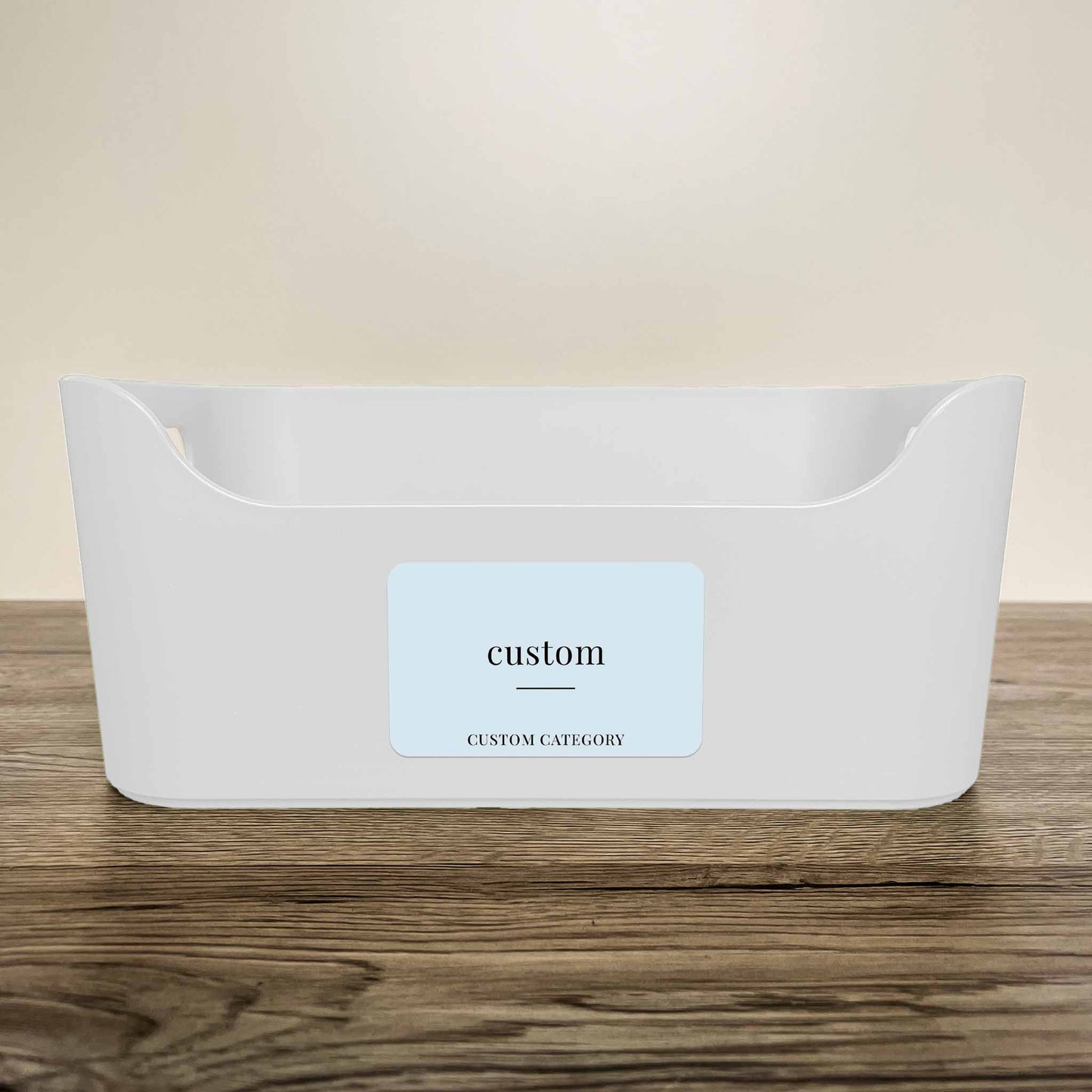 A white plastic storage basket with a customisable minimalist label in soft blue colour on the front, displayed on a wooden surface against a neutral beige background.