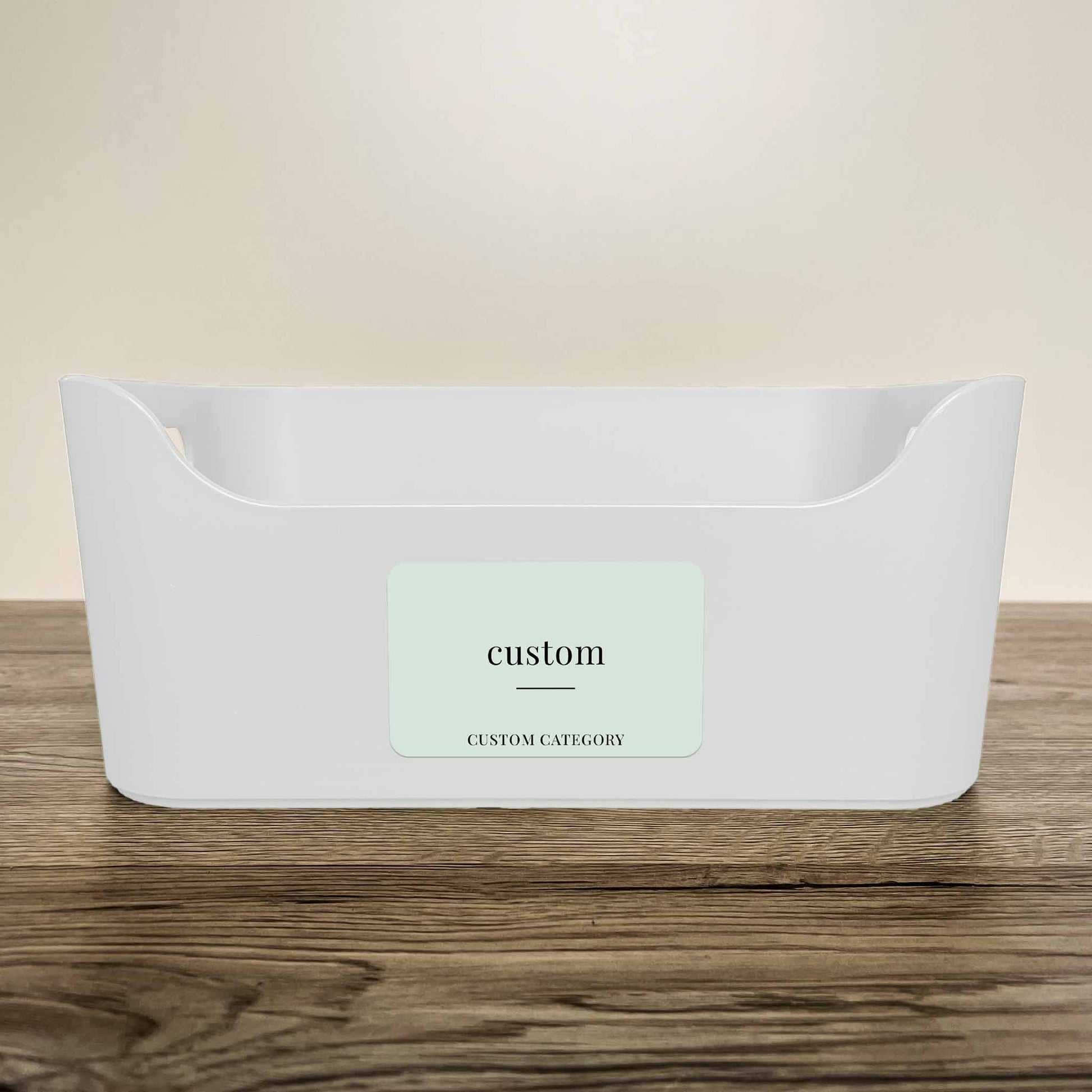 A white plastic storage basket with a customisable minimalist label in soft green colour on the front, displayed on a wooden surface against a neutral beige background.