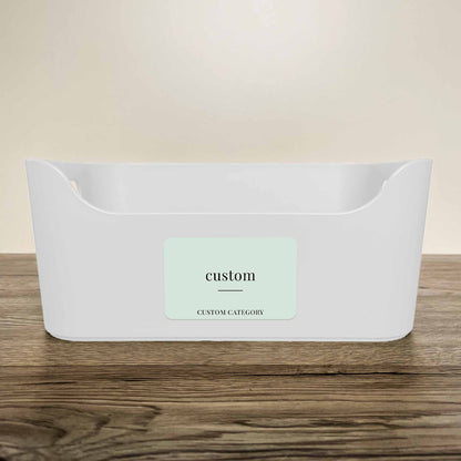 A white plastic storage basket with a customisable minimalist label in soft green colour on the front, displayed on a wooden surface against a neutral beige background.