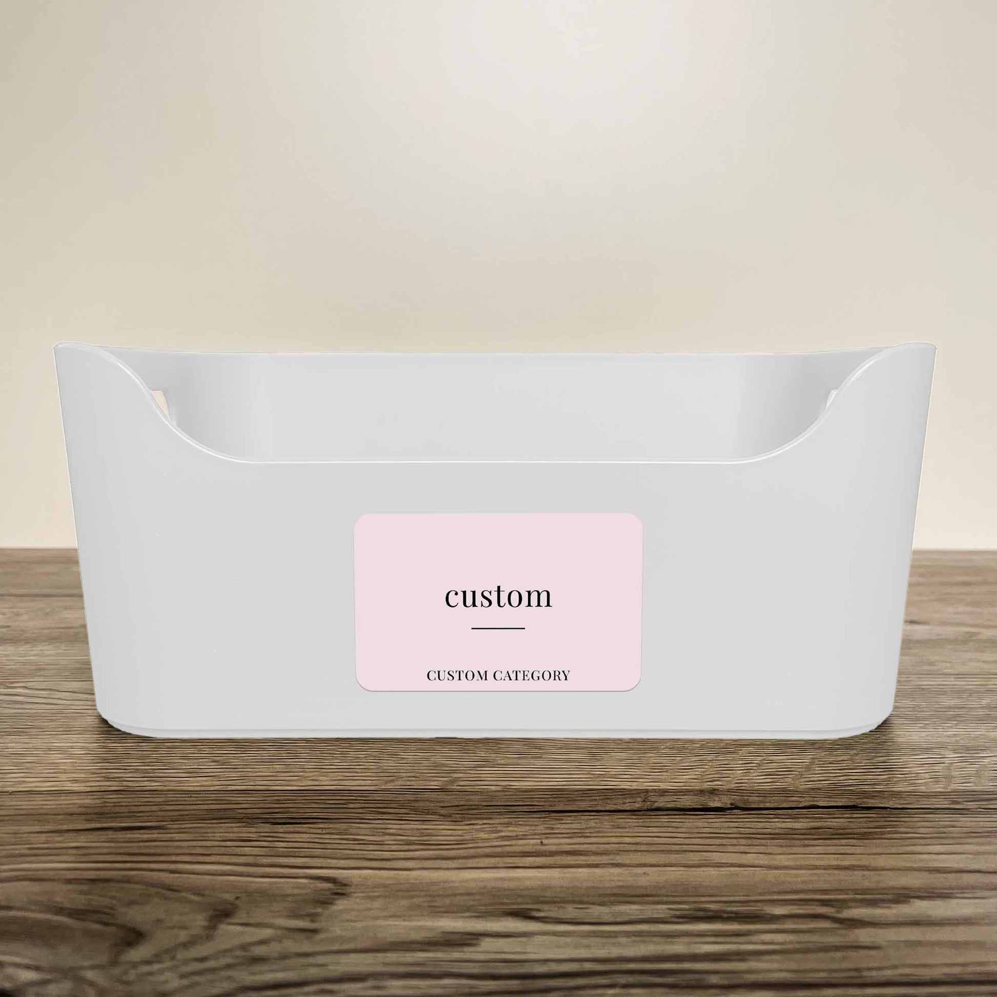 A white plastic storage basket with a customisable minimalist label in soft pink colour on the front, displayed on a wooden surface against a neutral beige background.