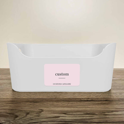 A white plastic storage basket with a customisable minimalist label in soft pink colour on the front, displayed on a wooden surface against a neutral beige background.