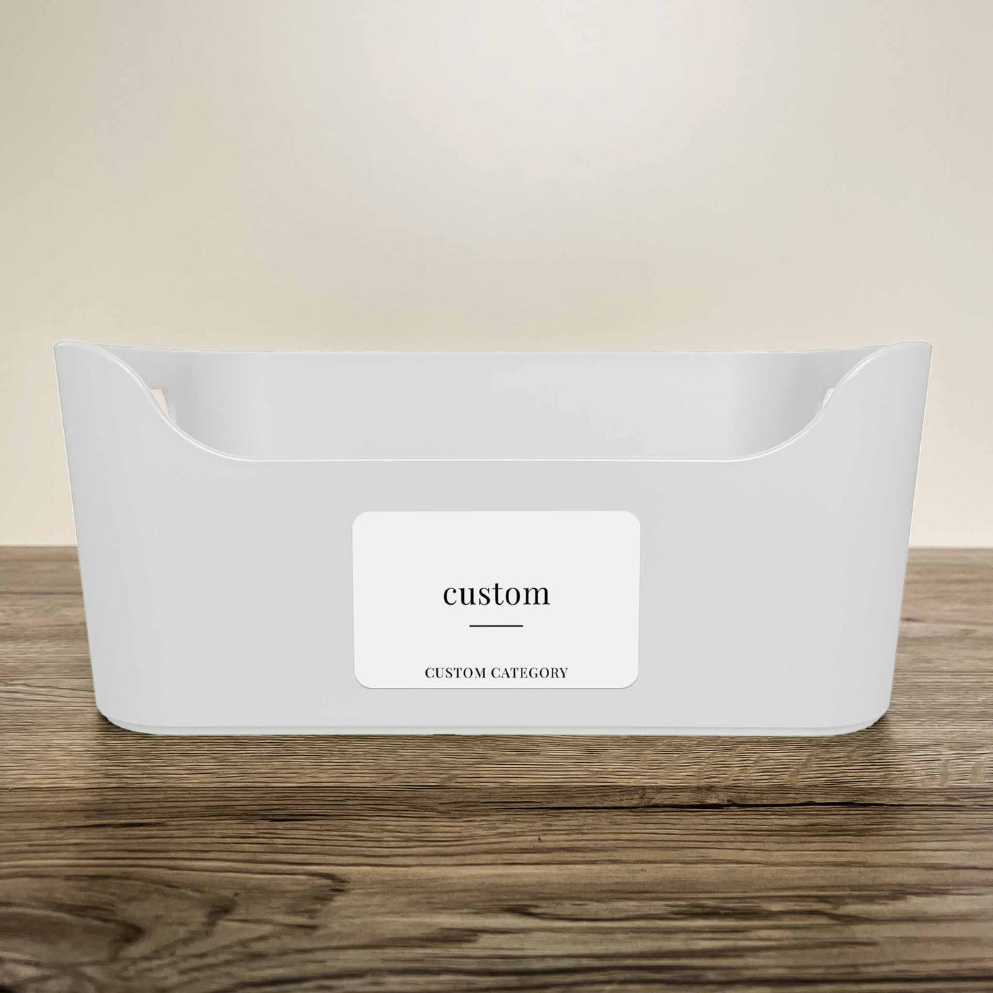 A white plastic storage basket with a customisable minimalist label in white colour on the front, displayed on a wooden surface against a neutral beige background.