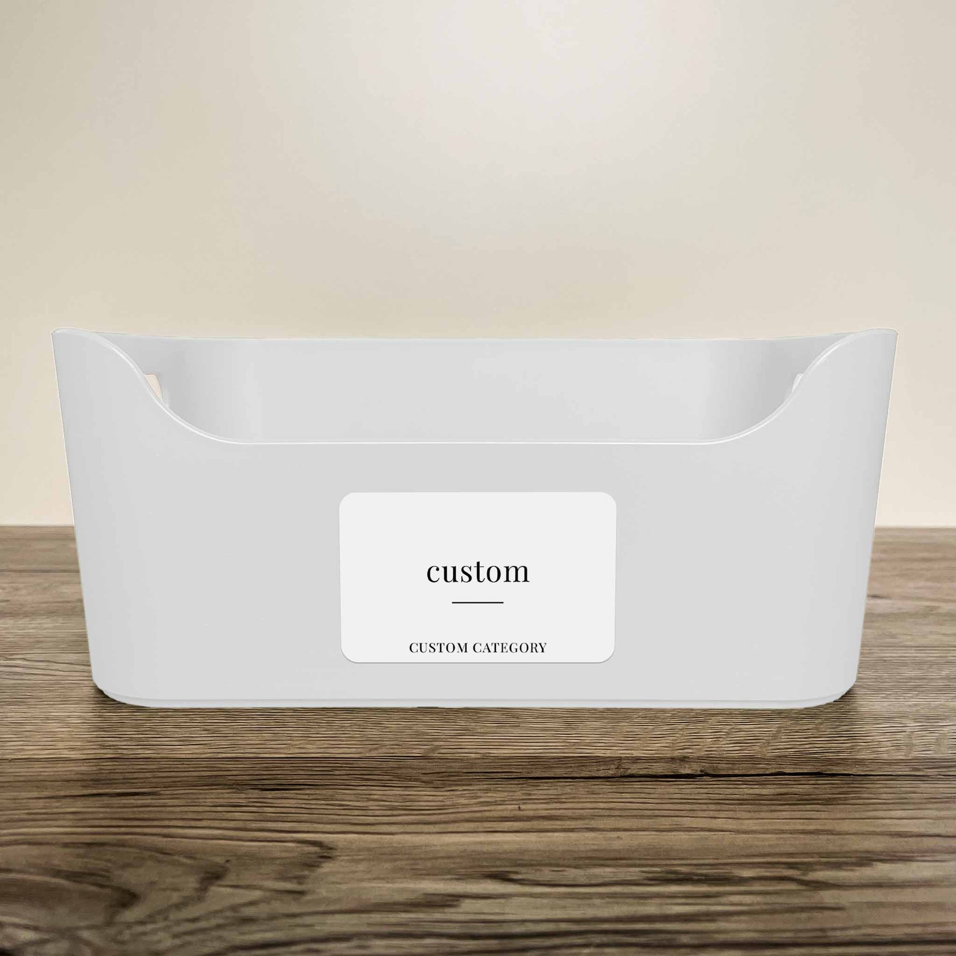 A white plastic storage basket with a customisable minimalist label in white colour on the front, displayed on a wooden surface against a neutral beige background.