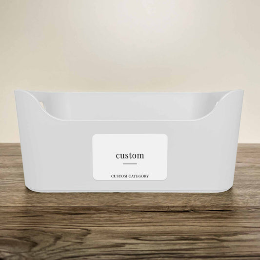 A white plastic storage basket with a customisable minimalist label in white colour on the front, displayed on a wooden surface against a neutral beige background.