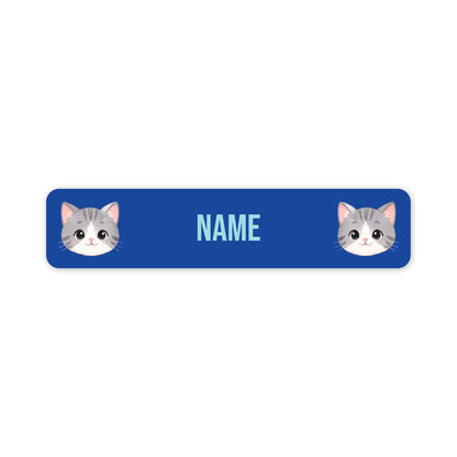 A blue rectangular name tag with rounded corners, featuring two cartoon gray-and-white cat faces at the left and right edges and the word 'NAME' in light blue in the centre, shown on a white background.