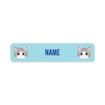 A horizontal light blue personalised name label with rounded corners, featuring two cartoon gray-and-white cat faces at the ends and the word 'NAME' in blue in the centre.