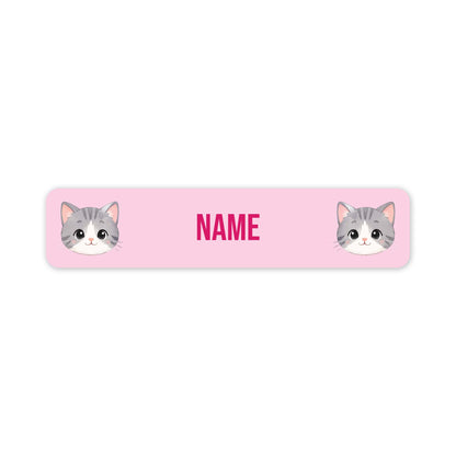 A pink rectangular label with rounded corners, featuring two cartoon gray-and-white cat faces at the left and right edges and the word 'NAME' in pink in the centre, shown on a white background.