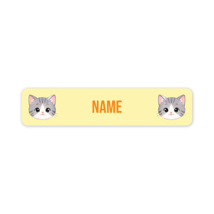 A rectangular pale yellow name tag with rounded corners, featuring two cartoon gray-and-white cat faces at the left and right edges and the word "NAME" in orange in the centre, shown on a white background.