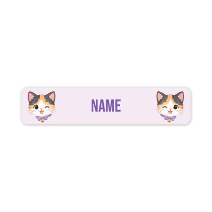 A horizontal light pink personalised name label with rounded corners, featuring two cartoon calico cat faces on the left and right sides and the word 'NAME' in purple in the centre.