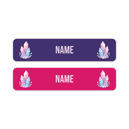 Two horizontal rectangular name tags on a white background, one dark purple and one bright pink, each with rounded corners, customisable 'NAME' text in the centre, and a pastel crystal illustration near the left and right edges.