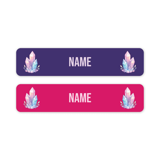 Two horizontal rectangular name tags on a white background, one dark purple and one bright pink, each with rounded corners, customisable 'NAME' text in the centre, and a pastel crystal illustration near the left and right edges.