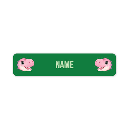 A green rectangular nameplate or sticker with rounded corners, featuring two pink cartoon dinosaur faces at the left and right edges and the word 'NAME' in the center for personalization.