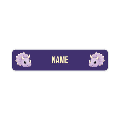 A horizontal purple personalized name label with rounded corners, featuring two cute light purple cartoon triceratops dinosaur faces on the left and right sides and the word 'NAME' in the center in a pale yellow font.