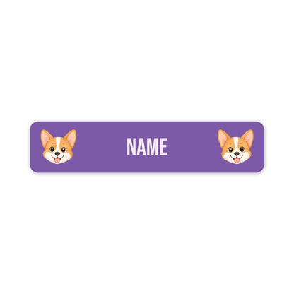 A purple rectangular name tag with rounded corners, featuring two cartoon corgi faces on the left and right sides and the word 'NAME' in white centered in the middle, shown on a plain white background.