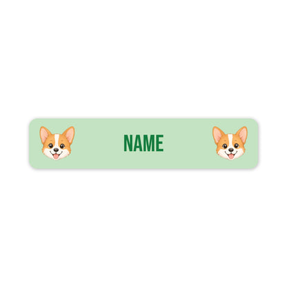 A horizontal light green personalized name label with rounded corners, featuring two cartoon corgi faces and the text 'NAME' in green in the center, shown on a plain white background.