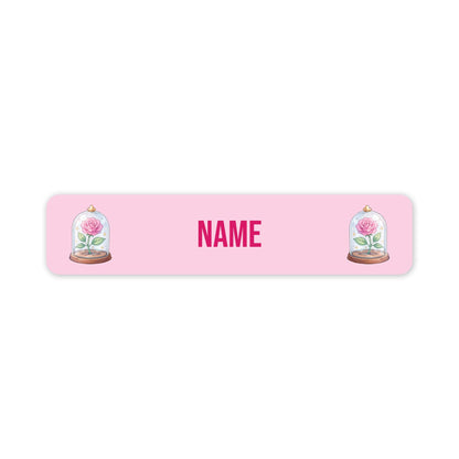A pink rectangular personalized name plaque with rounded corners, featuring the word 'NAME' in bold pink letters in the centre and an illustration of a pink rose under a glass cloche on each side.