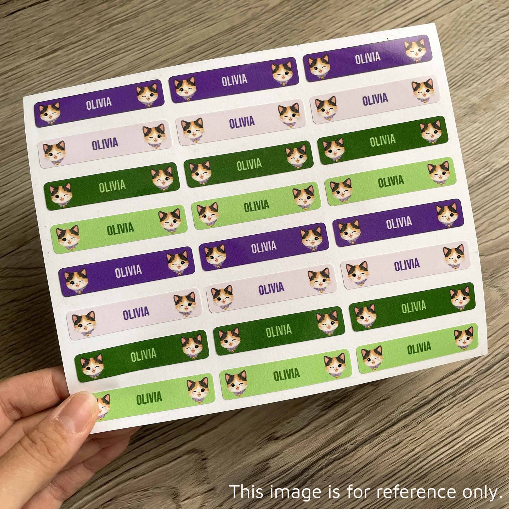 A hand holds a sheet of rectangular stickers printed with the name 'OLIVIA' repeated in white text over purple, pink, green, and dark green backgrounds, with small cat face illustrations. The sheet is photographed on a wood surface.