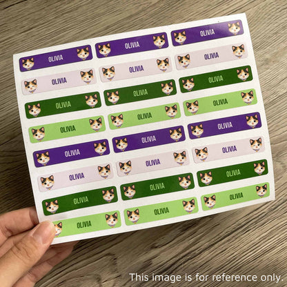 A hand holds a sheet of rectangular stickers printed with the name 'OLIVIA' repeated in white text over purple, pink, green, and dark green backgrounds, with small cat face illustrations. The sheet is photographed on a wood surface.