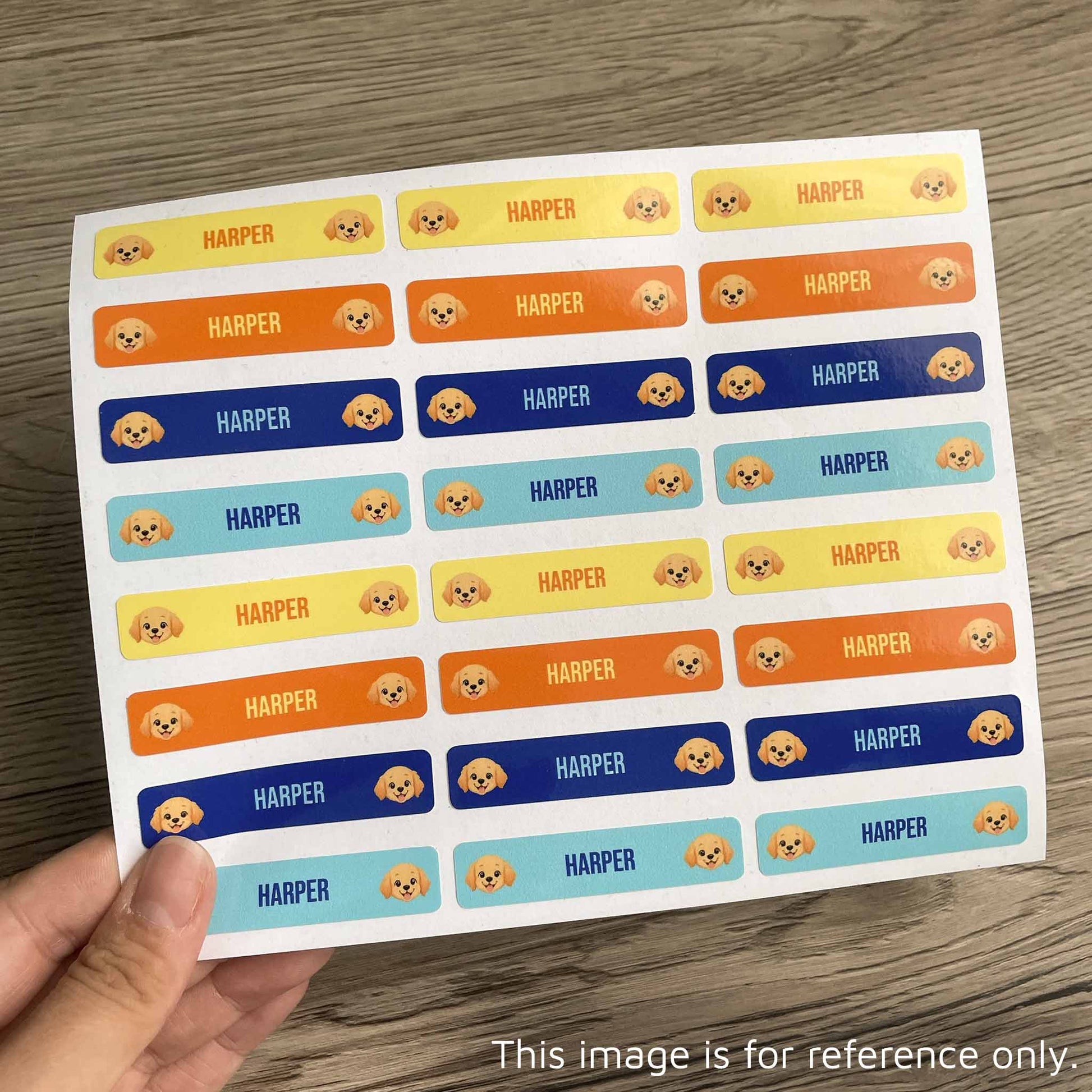 A hand holds a sheet of rectangular stickers on a wood surface. Each sticker has a small cartoon dog face and the name HARPER in different color blocks: yellow, orange, blue, and light blue.