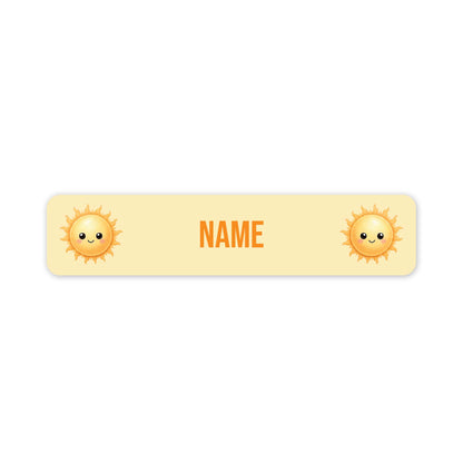 A horizontal pale yellow name tag with rounded corners, featuring two cute smiling sun illustrations on the left and right sides and the customisable word 'NAME' in orange centered in the middle, shown on a white background.