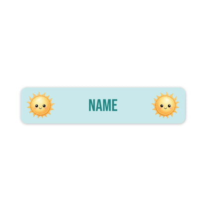 A horizontal light blue personalised name label with rounded corners, featuring two cute yellow smiling sun icons on the left and right sides and the customisable word 'NAME' in teal in the centre, shown on a plain white background.